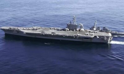 Large aircraft carrier at sea with fighter jets lined up on the deck and a smaller escort ship nearby in blue water.