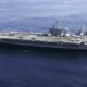 Large aircraft carrier at sea with fighter jets lined up on the deck and a smaller escort ship nearby in blue water.