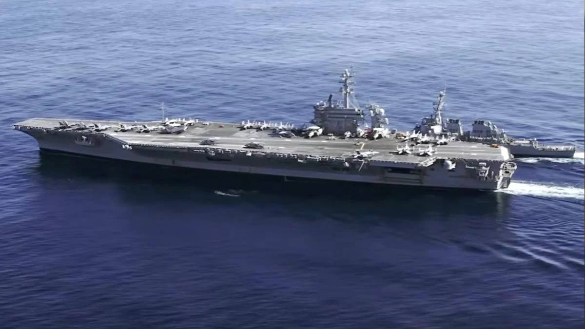 Large aircraft carrier at sea with fighter jets lined up on the deck and a smaller escort ship nearby in blue water.