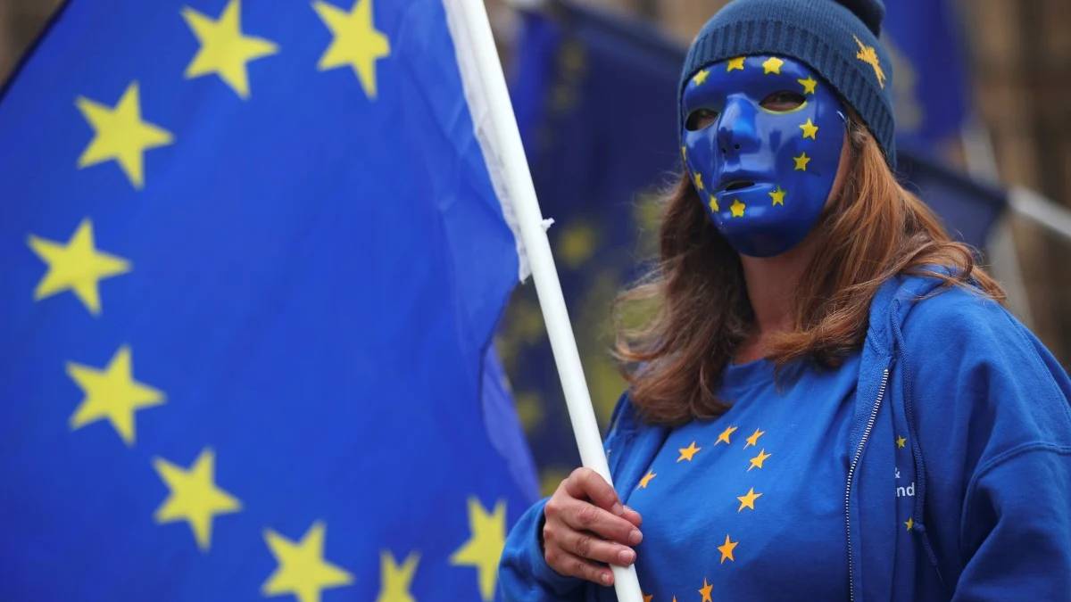 Protester wearing a blue EU flag face mask and shirt, holding a white flagpole with a large EU flag in the background.