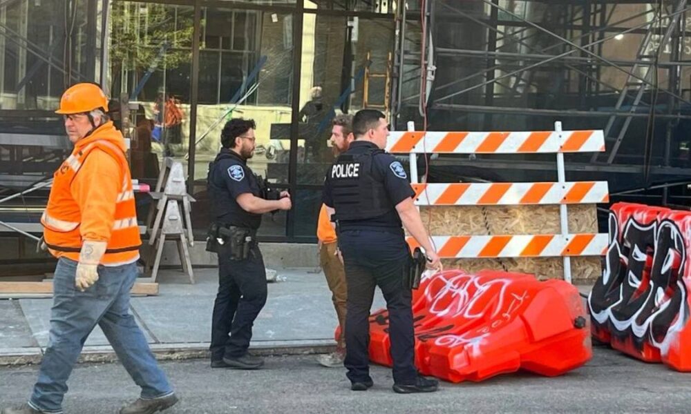 Construction worker in an orange safety vest and hard hat walks past police officers near orange-and-white barricades outside a building under construction.