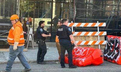 Construction worker in an orange safety vest and hard hat walks past police officers near orange-and-white barricades outside a building under construction.