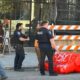 Construction worker in an orange safety vest and hard hat walks past police officers near orange-and-white barricades outside a building under construction.
