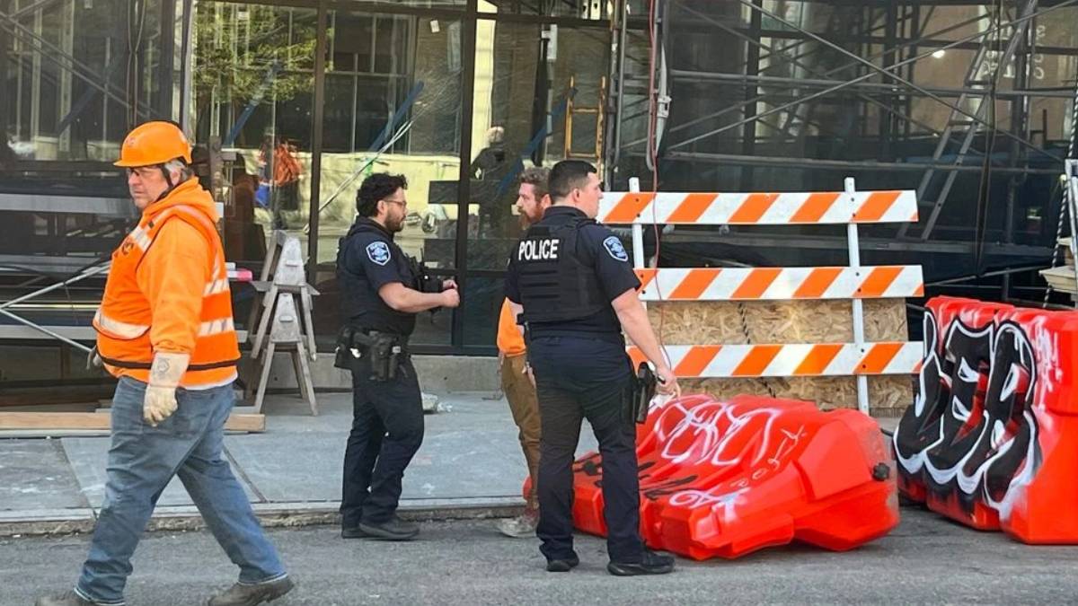 Construction worker in an orange safety vest and hard hat walks past police officers near orange-and-white barricades outside a building under construction.