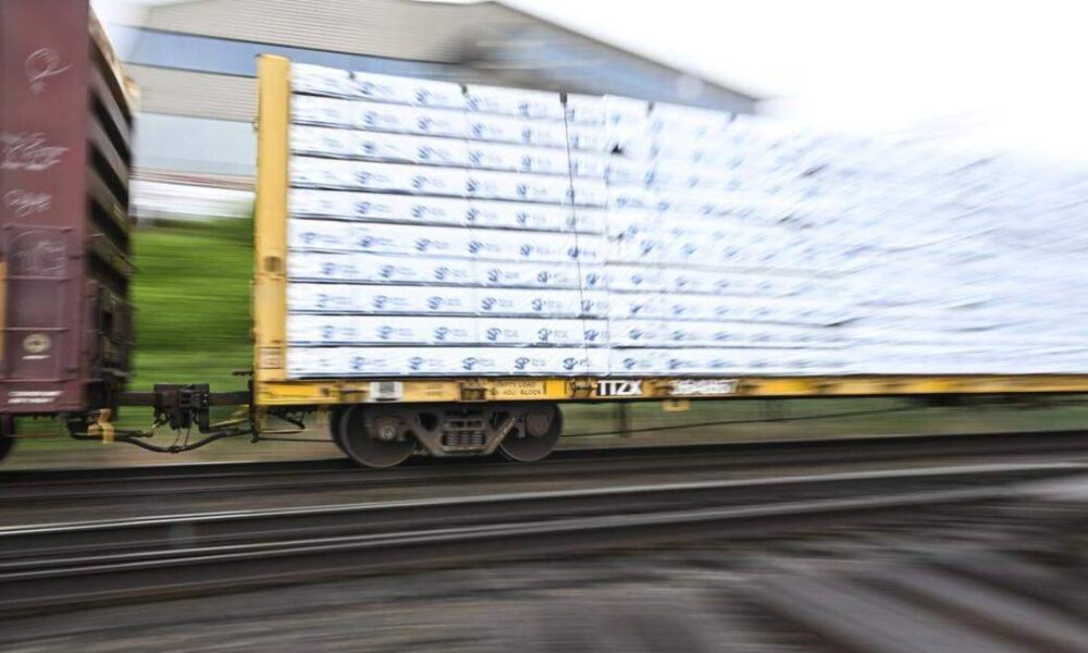 Blurry cargo train car on a flatbed carrying stacked white-wrapped pallets, in motion.