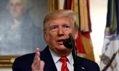 Donald Trump speaking into a microphone, gesturing with his hand in a dark suit and red tie.