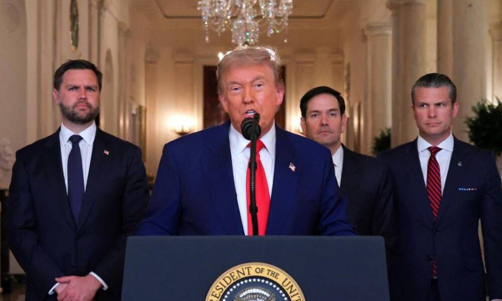 Donald Trump speaks at a lectern with the Presidential seal, flanked by three suited aides in a hallway setting.