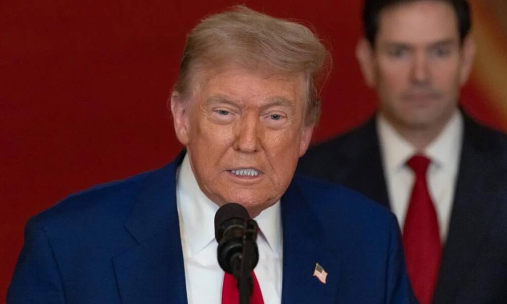 Donald Trump delivering remarks into a microphone, wearing a blue suit and red tie, with a suited man in the background.