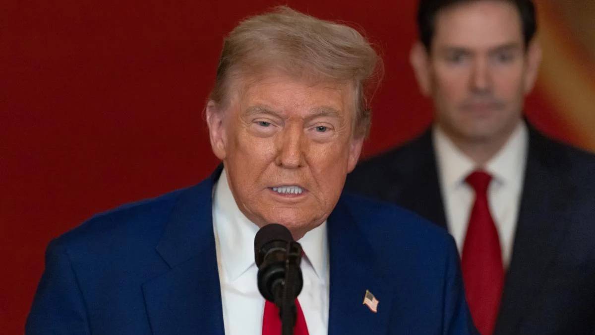 Donald Trump delivering remarks into a microphone, wearing a blue suit and red tie, with a suited man in the background.
