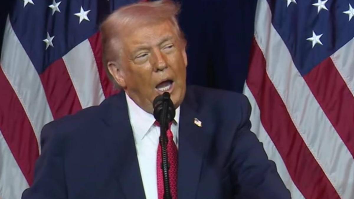 Man in a dark suit and red tie speaks into a microphone with U.S. flags behind him.