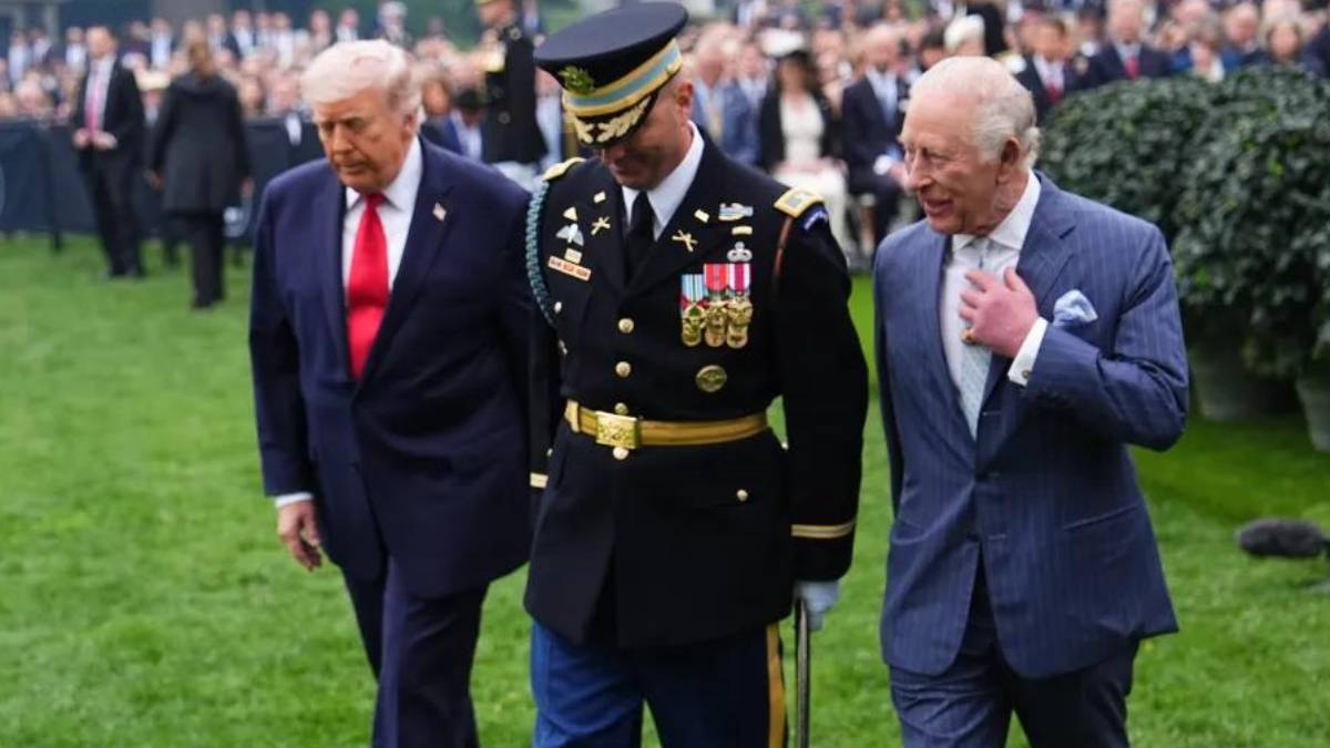 Three men in suits and a decorated military officer walk across a grassy lawn during a formal event, with a crowd in the background.
