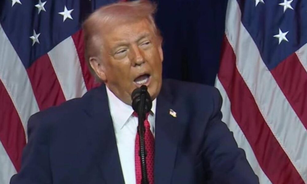 A man in a dark suit and red tie speaks at a podium with U.S. flags in the background, microphone in front of him.