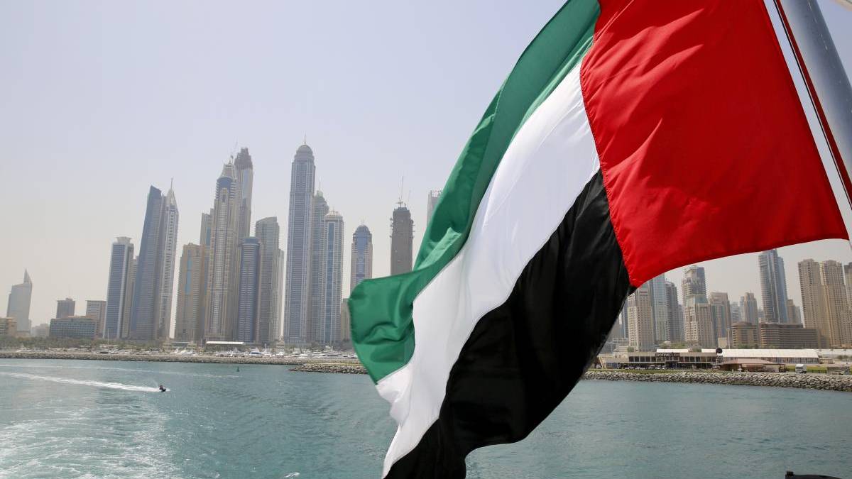 Flag of the United Arab Emirates waving in front of Dubai's skyline over the water.