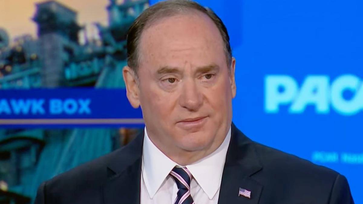 Man in a dark suit and striped tie on a television set, wearing an American flag lapel pin and a serious expression.