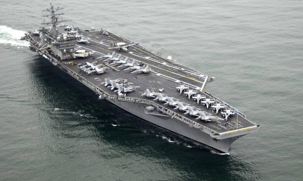 Military aircraft carrier at sea with multiple fighter jets lined up on deck and crew areas visible on the flight deckwise.