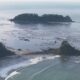 Aerial view of a rocky coastal shoreline with a small forested island in calm ocean waters behind.