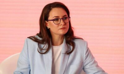 Woman with glasses and brown hair in a light blue blazer sits on a panel against a pink backdrop, looking to the side.