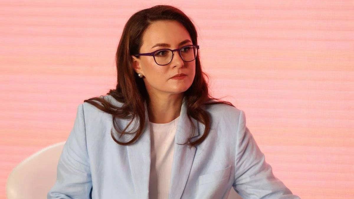 Woman with glasses and brown hair in a light blue blazer sits on a panel against a pink backdrop, looking to the side.