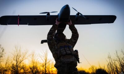 Soldier in camouflage gear lifts a small propeller plane overhead at sunset.