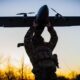 Soldier in camouflage gear lifts a small propeller plane overhead at sunset.