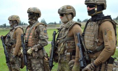 Four soldiers in camouflage combat gear stand in a grassy field, each holding a rifle and wearing helmets, masks, and tactical vests.