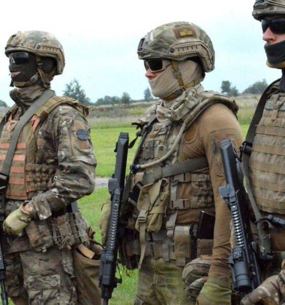 Four soldiers in camouflage combat gear stand in a grassy field, each holding a rifle and wearing helmets, masks, and tactical vests.