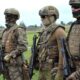Four soldiers in camouflage combat gear stand in a grassy field, each holding a rifle and wearing helmets, masks, and tactical vests.