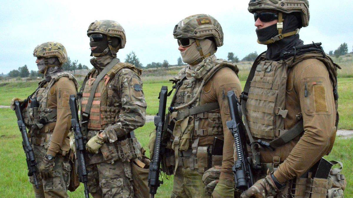 Four soldiers in camouflage combat gear stand in a grassy field, each holding a rifle and wearing helmets, masks, and tactical vests.