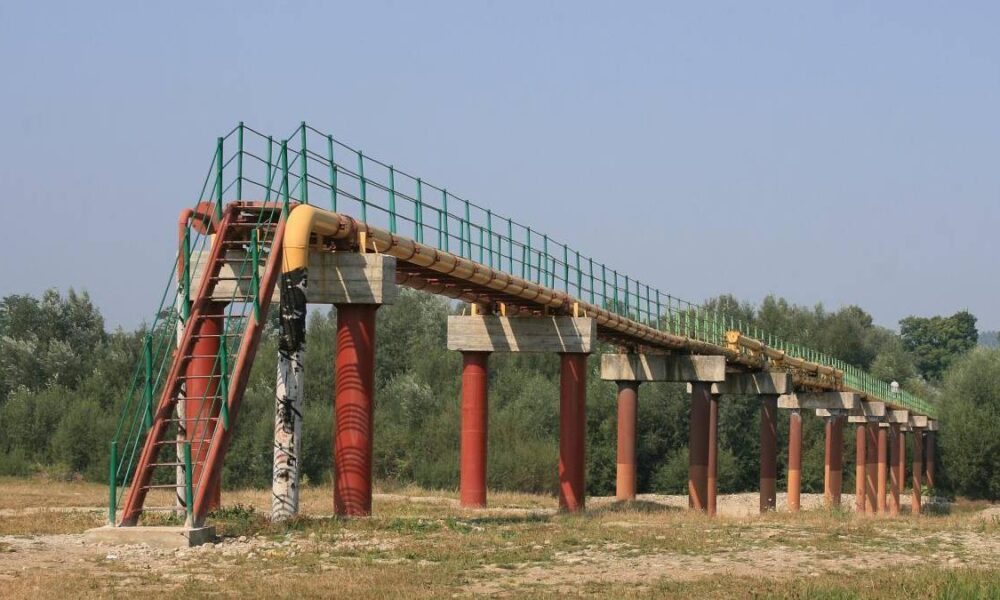 Elevated pipeline on red columns with green railing, accessible by a red stairway, in a rural field.
