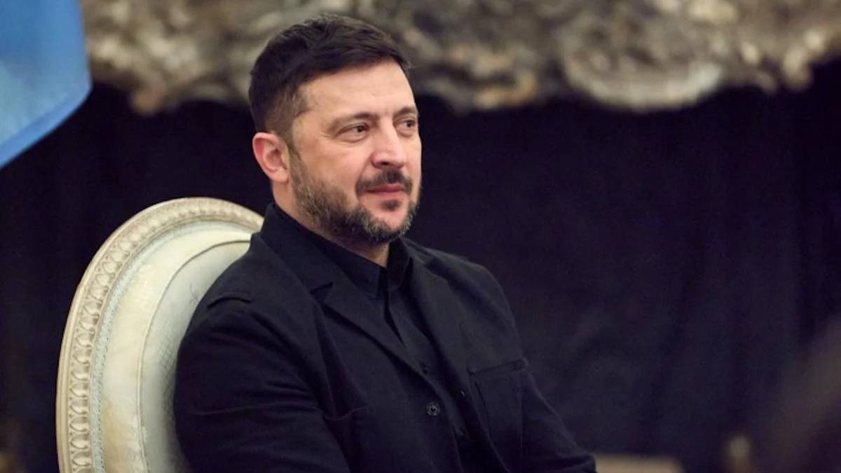 Man with short dark hair and beard sits in a black suit on an ornate light chair, looking to the side.