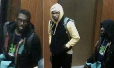 Three young men in hoodies and jackets stand indoors by a wooden wall, backpacks on, looking toward the camera (security-style footage).