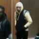 Three young men in hoodies and jackets stand indoors by a wooden wall, backpacks on, looking toward the camera (security-style footage).