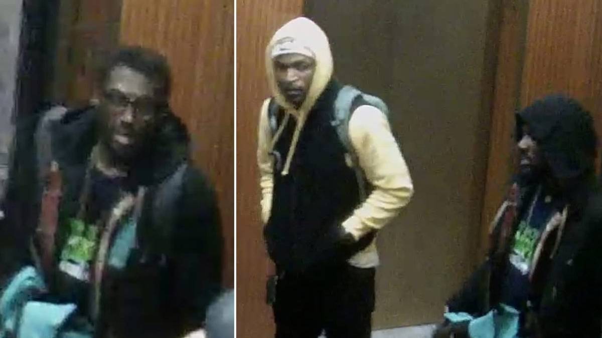 Three young men in hoodies and jackets stand indoors by a wooden wall, backpacks on, looking toward the camera (security-style footage).