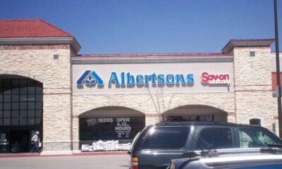Albertsons storefront with blue sign and Savon Pharmacy to the right, brick building and cars in a shopping center parking lot under a clear blue sky