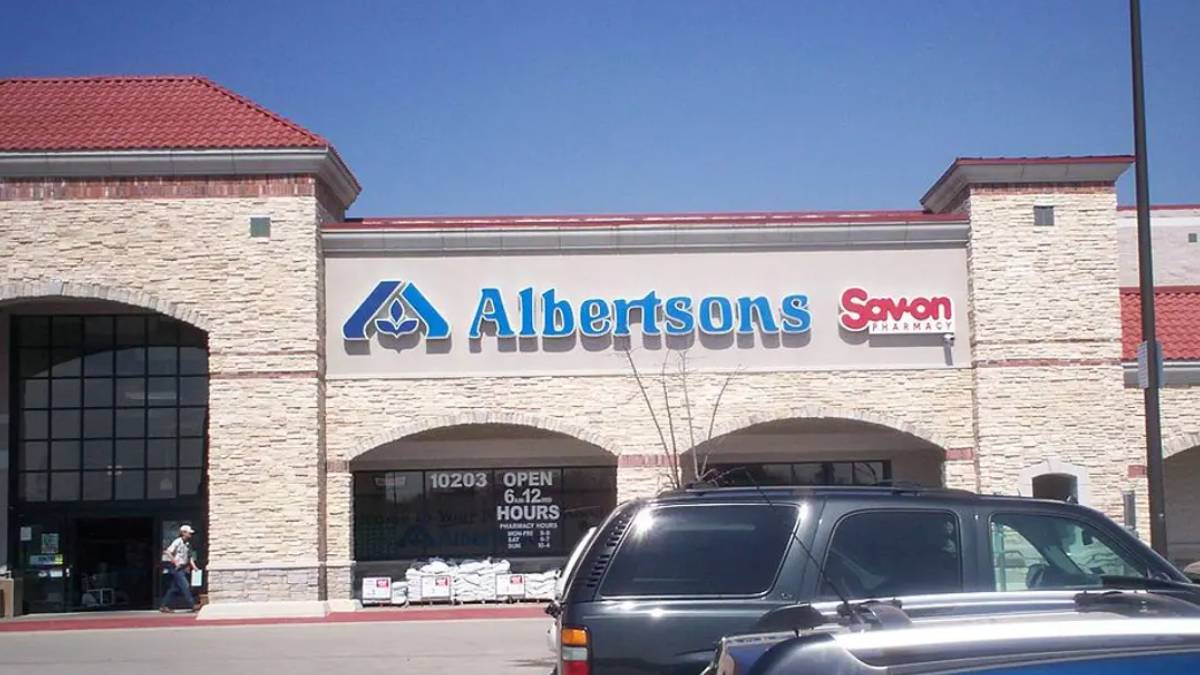Albertsons storefront with blue sign and Savon Pharmacy to the right, brick building and cars in a shopping center parking lot under a clear blue sky