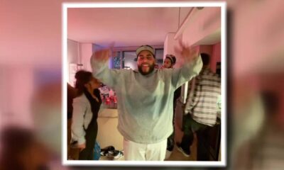 Smiling man in a gray sweatshirt and cap raises both arms in a crowded indoor party room.