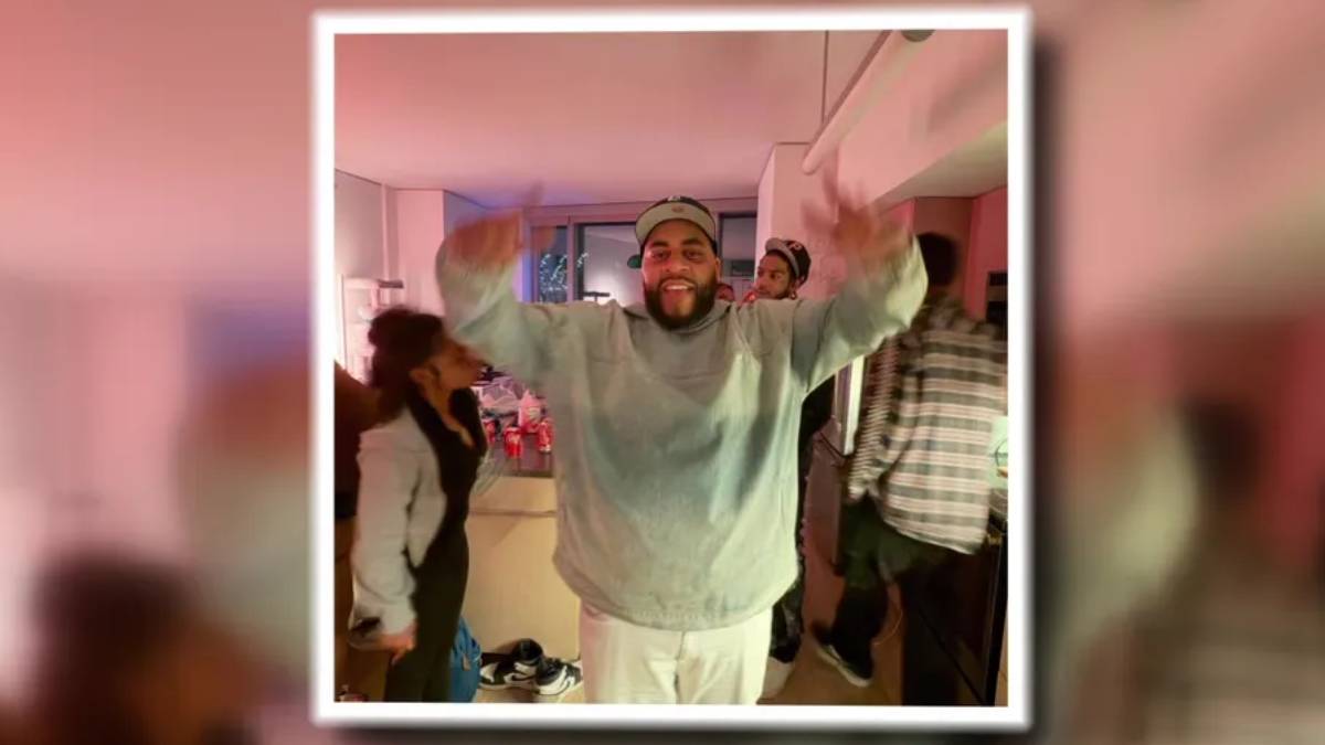 Smiling man in a gray sweatshirt and cap raises both arms in a crowded indoor party room.
