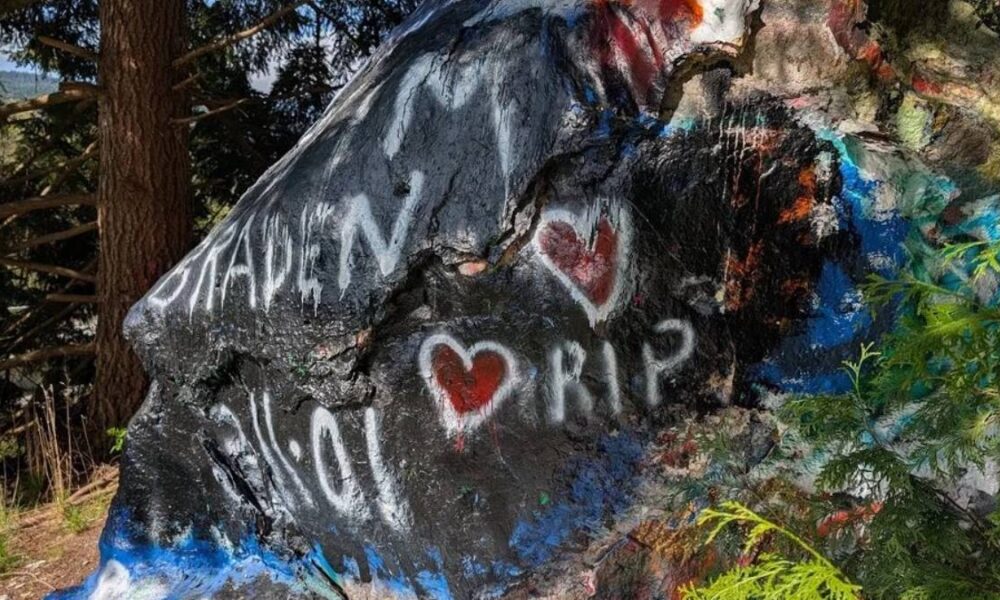 Rock with colorful graffiti on a mountainside: white hearts and the words 'RIP' painted among other colors, with trees in the background.