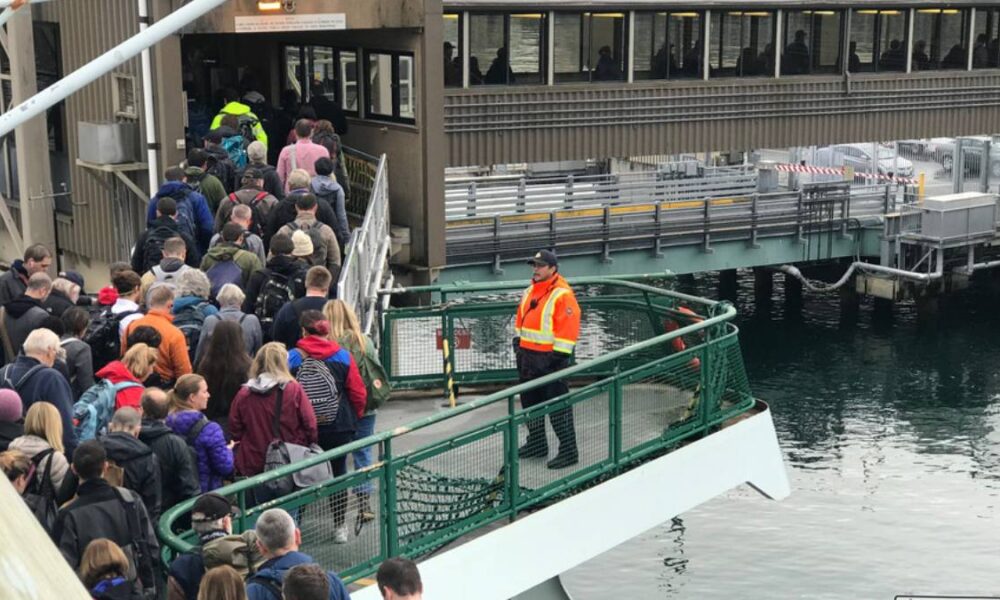 Crowd of passengers waiting in line on a boarding ramp toward a ferry at a dock, with a uniformed staff member in an orange vest nearby.