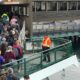 Crowd of passengers waiting in line on a boarding ramp toward a ferry at a dock, with a uniformed staff member in an orange vest nearby.