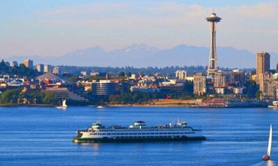 Seattle skyline across the water with the Space Needle, a large white ferry in the foreground, and a small sailboat nearby on a calm blue bay.