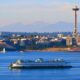 Seattle skyline across the water with the Space Needle, a large white ferry in the foreground, and a small sailboat nearby on a calm blue bay.