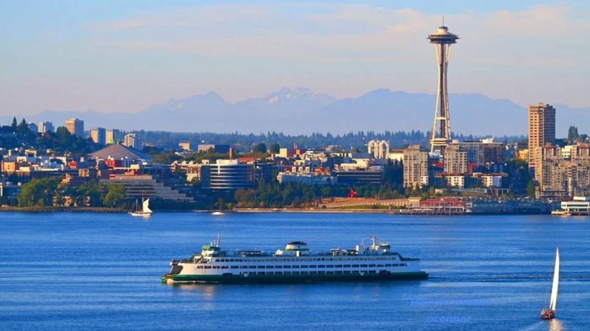 Seattle skyline across the water with the Space Needle, a large white ferry in the foreground, and a small sailboat nearby on a calm blue bay.