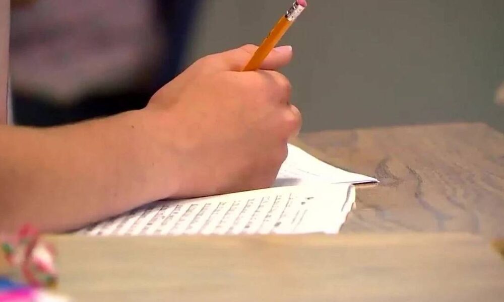Close-up of a hand writing with a yellow pencil on lined paper at a wooden desk.