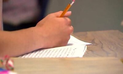 Close-up of a hand writing with a yellow pencil on lined paper at a wooden desk.