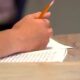 Close-up of a hand writing with a yellow pencil on lined paper at a wooden desk.