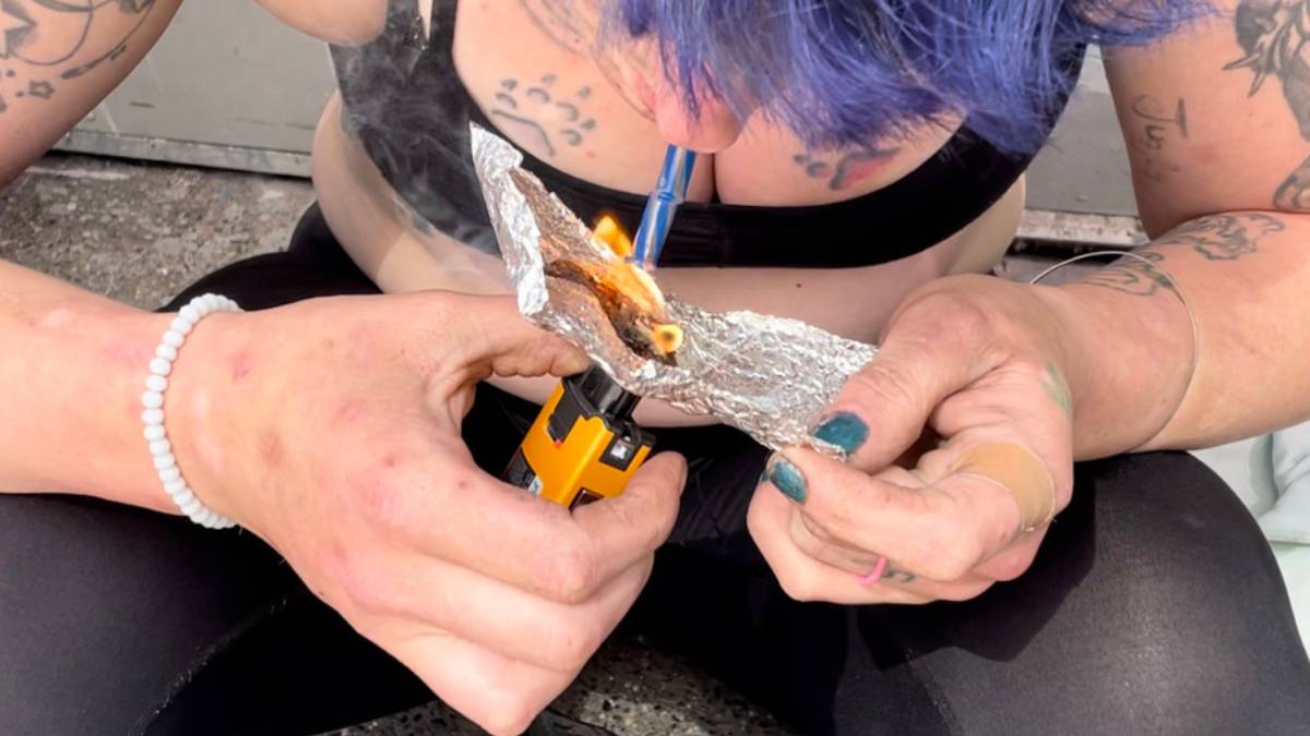 Close-up of hands heating a substance on aluminum foil with a small torch, the person has blue hair and visible tattoos.
