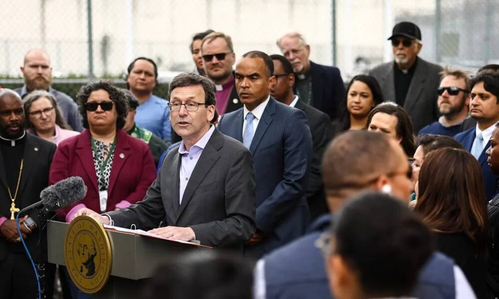 Politician at a podium giving a press conference, surrounded by a diverse crowd of onlookers and reporters.