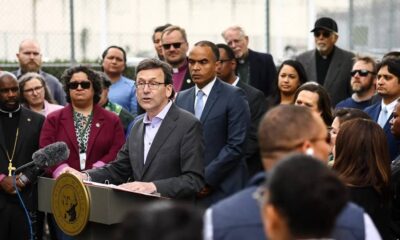 Politician at a podium giving a press conference, surrounded by a diverse crowd of onlookers and reporters.