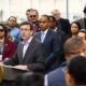 Politician at a podium giving a press conference, surrounded by a diverse crowd of onlookers and reporters.